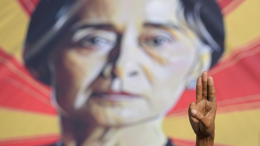 A person shows the three-fingers salute in front of a placard with the image of Aung San Suu Kyi.