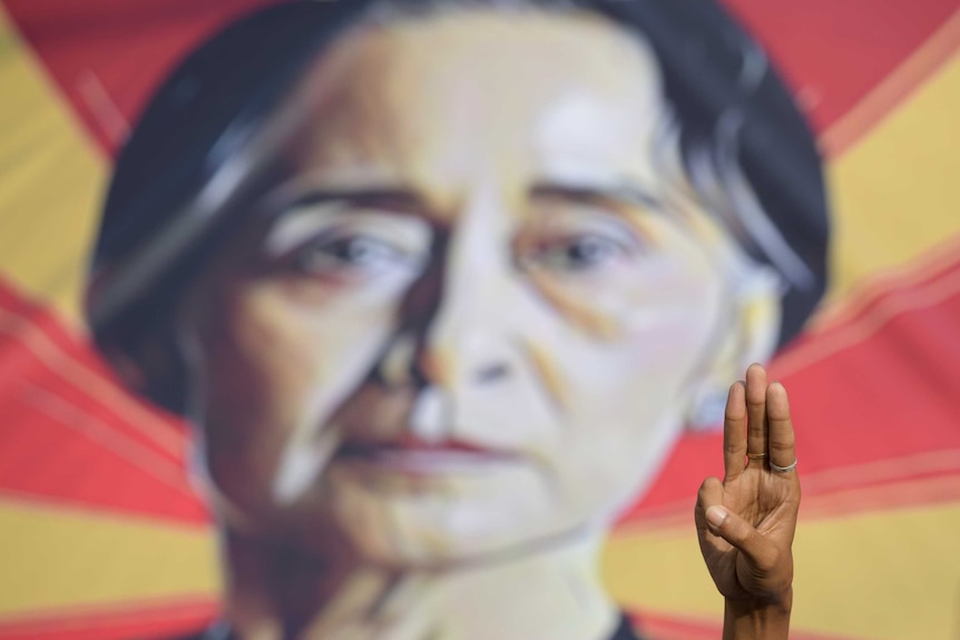 A person shows the three-fingers salute in front of a placard with the image of Aung San Suu Kyi.