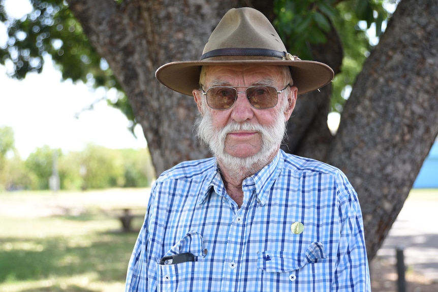Man with grey beard looks at the camera, wearing sunglasses and an akubra-style hate, under a tree