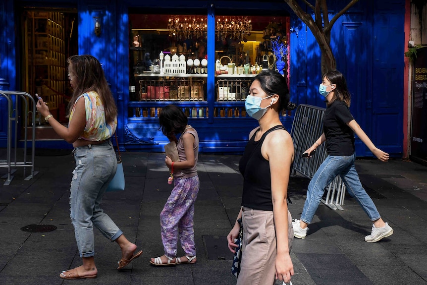 Several women wearing face masks walk along a shopping street. 