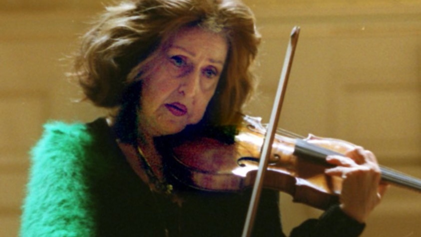 Violinist Ida Haendel plays on stage wearing a green jumper. She looks seriously into the distance.
