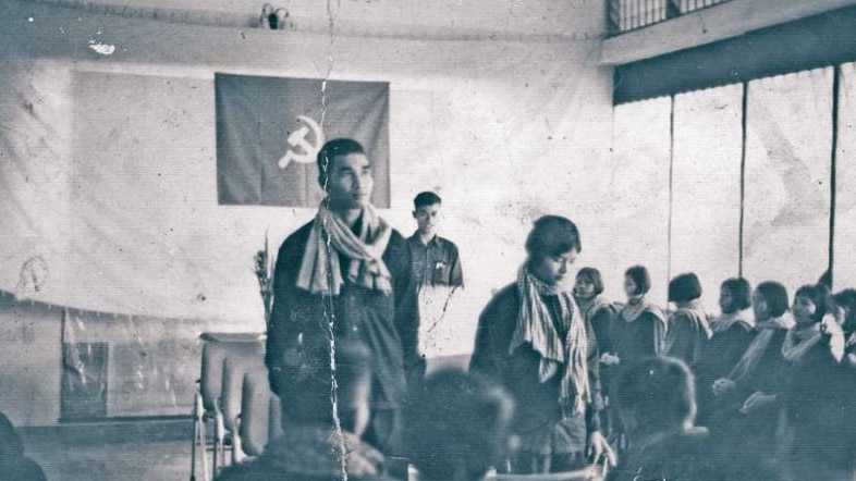 A black and white photo showing a man standing next to a woman with a hammer and sickle flag in the background.