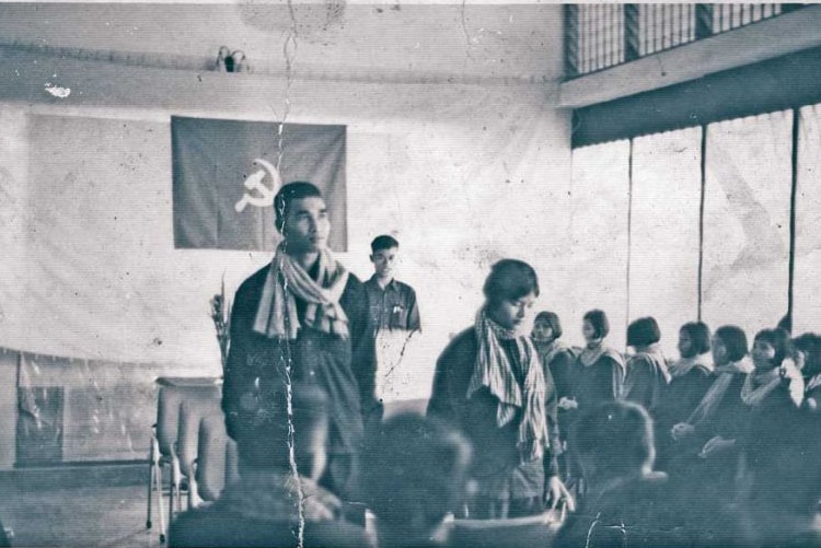 A black and white photo showing a man standing next to a woman with a hammer and sickle flag in the background.