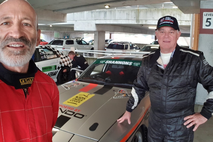 A balding man and a man in a cap stand in front of a rally car