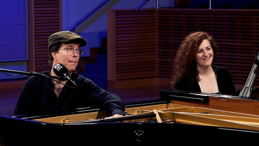 A man and a woman sit at two grand pianos. The man has his hand inside the open piano lid and the woman smiles.