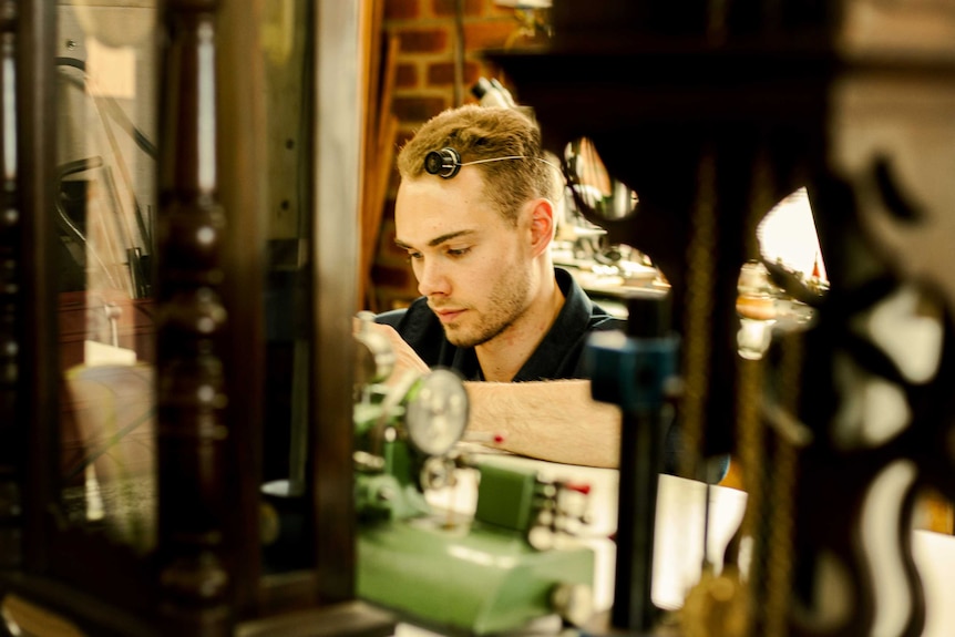 A young man wearing an eyepiece sits at a workshop desk, obscured by tall clocks.