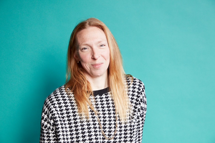 Portrait of choreographer Jo Lloyd, a middle-aged white woman sitting on a stool, teal backdrop, looking serious