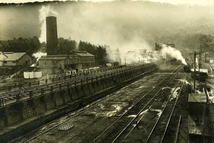 a black and white photo of train tracks with mining buildings behind 