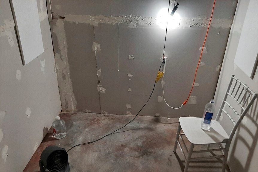Inside a cinder block cell with a chair, a light, unplastered wall, a fan and concreate ground. Very confined