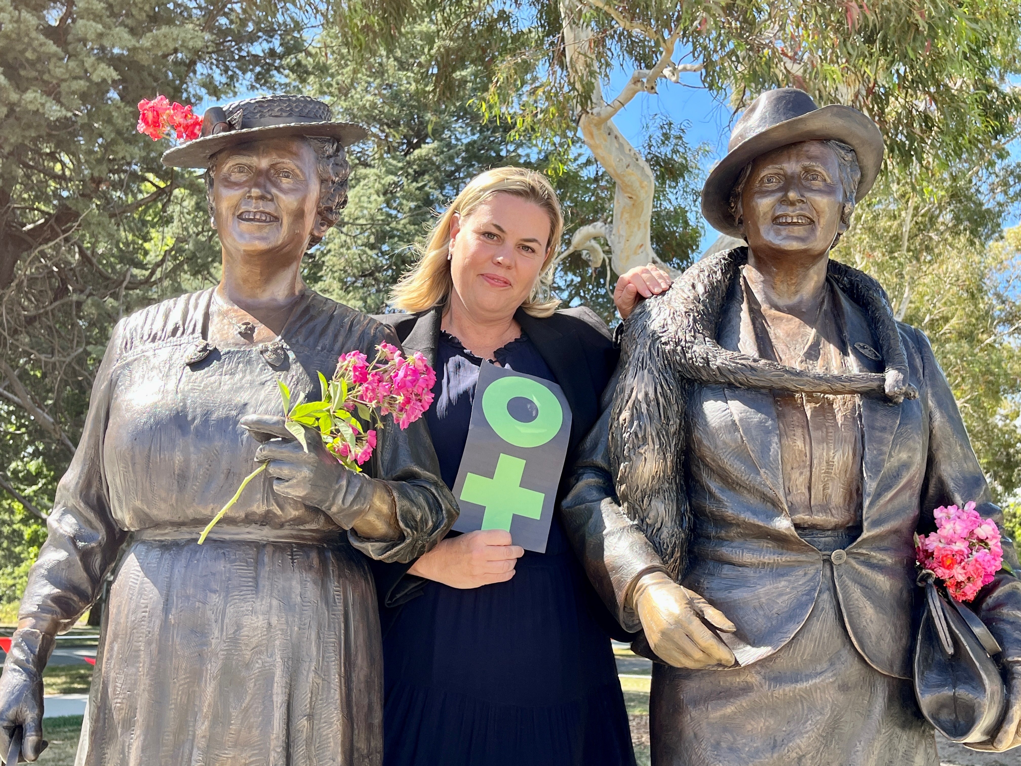 Middle-aged, blonde woman in dark blouse and blazer smiles at camera, standing in-between two bronze statues of women 