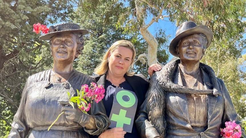 Middle-aged, blonde woman in dark blouse and blazer smiles at camera, standing in-between two bronze statues of women 