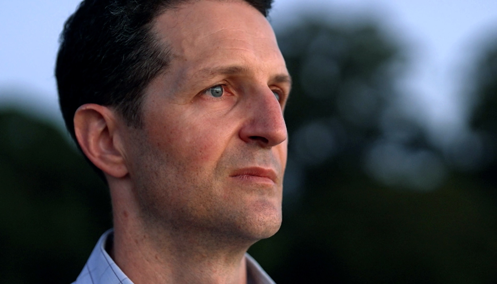 A close up of a man's face, he is standing outdoors looking straight ahead with a serious, slightly sad expression.