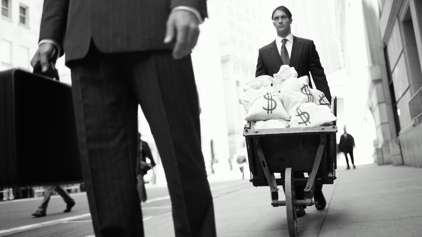 Two men in business suits walk down a city street, one pushing a wheelbarrow full of money. the other carries a briefcase
