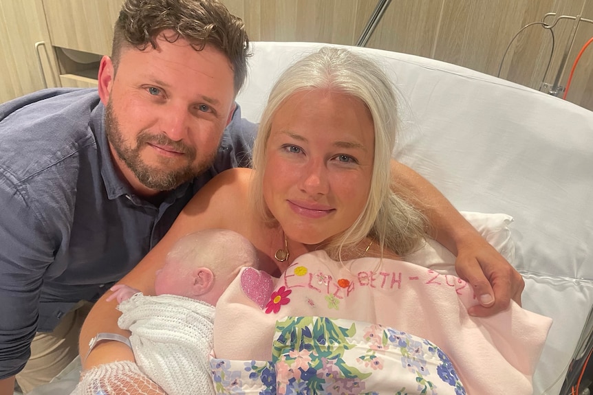 A woman with blonde hair poses for a photo with with her partner and baby in a hospital bed