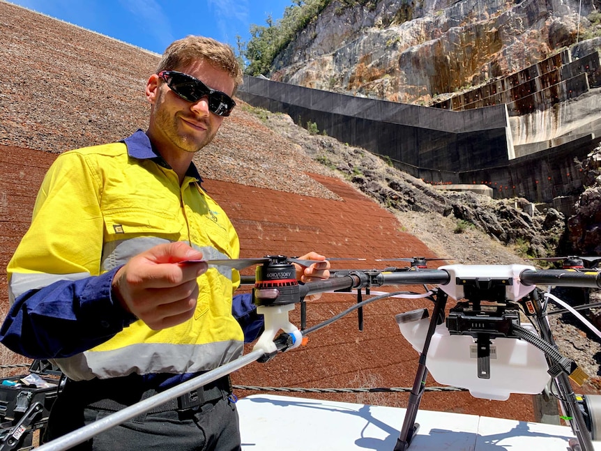 A man is wearing work gear and setting up his drone to fly and spray weeds in a dam.