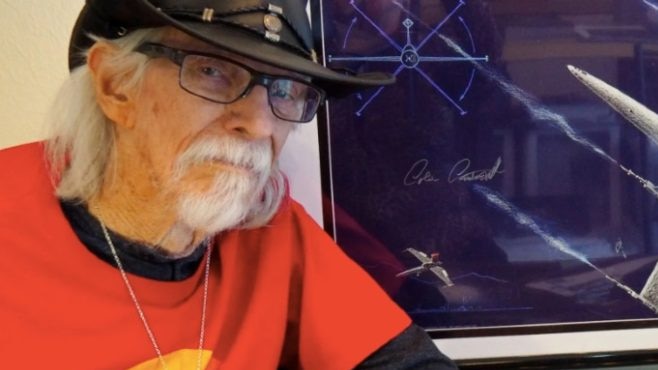 An older man with a short grey moustache wearing a leather black hat sits beside a picture of a spaceship 