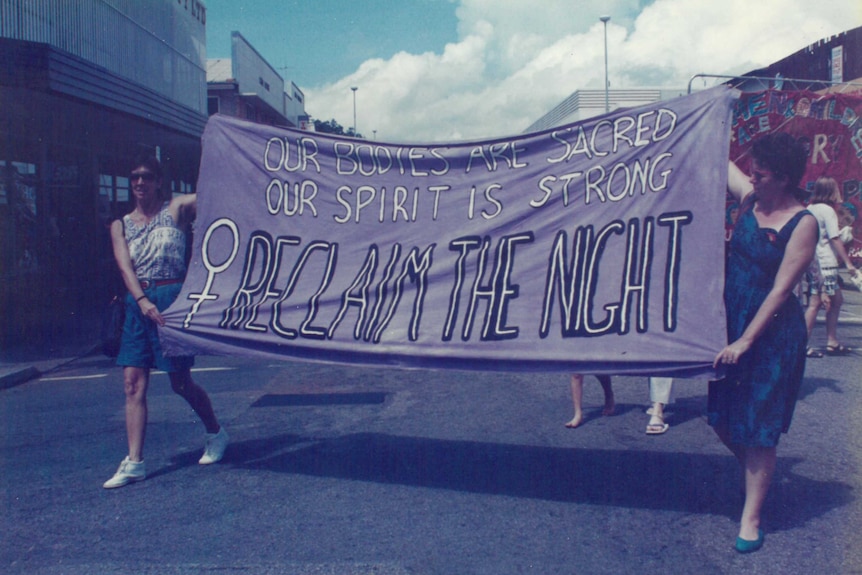 Two women march in Darwin's CBD holding a banner reading 'our bodies are sacred, our spirit is strong, reclaim the night'.