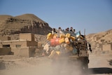 An overloaded truck drives through a village made of mudbricks set against hills.