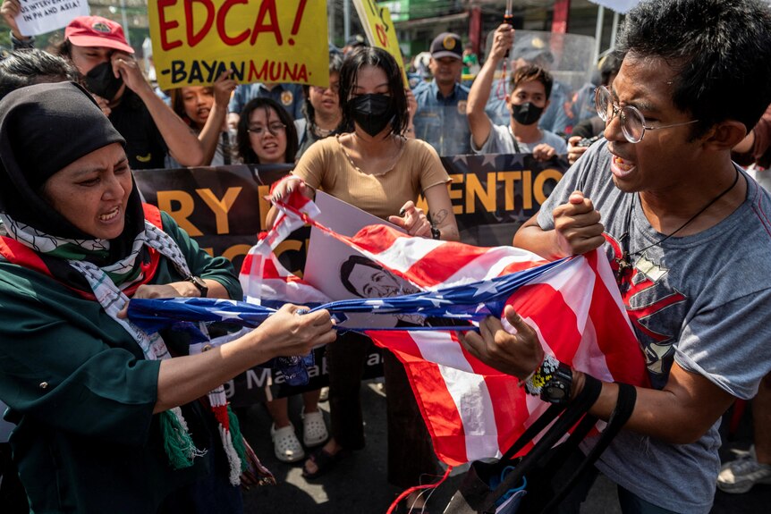 Filipino activists tear a US flag during a protest, with several holding placards in the background.