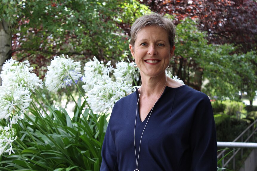 Woman with short light hair in navy blouse smiles at the camera. Agapanthus and trees are in the background.