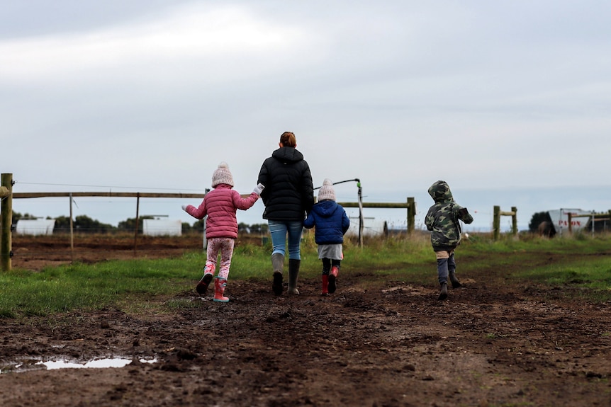 Mother and three young children wearing warm clothes walk on muddy path with paddocks either side