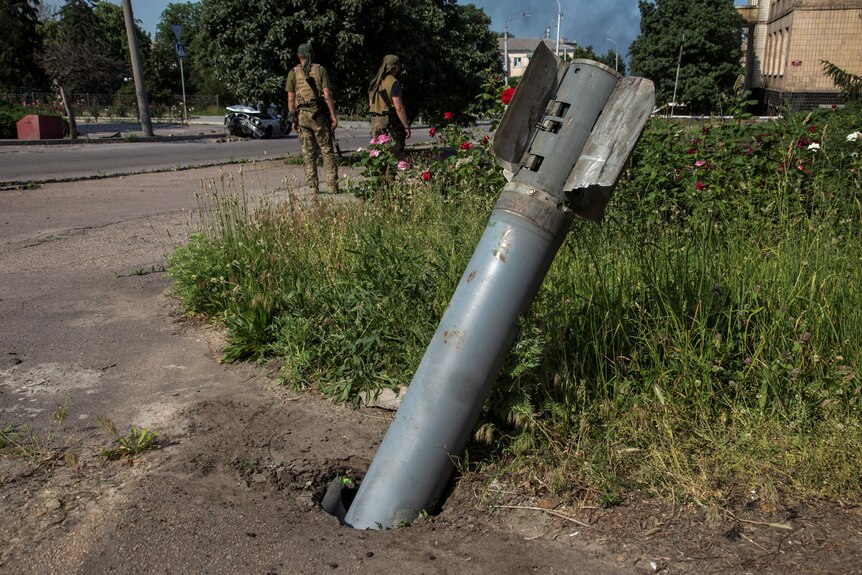 Two men walk past an unexploded shell stuck in the ground.