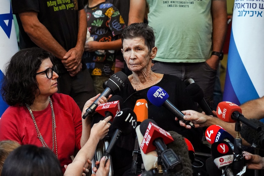 An elderly woman with short dark hair speaks in front of a crowd of journalists' microphones.
