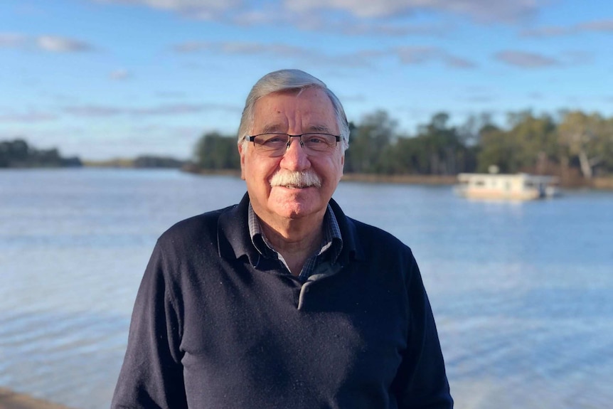 A man with white hair, a white moustache and glasses, dressed in a navy jumper, smiles at the camera on the river's edge.