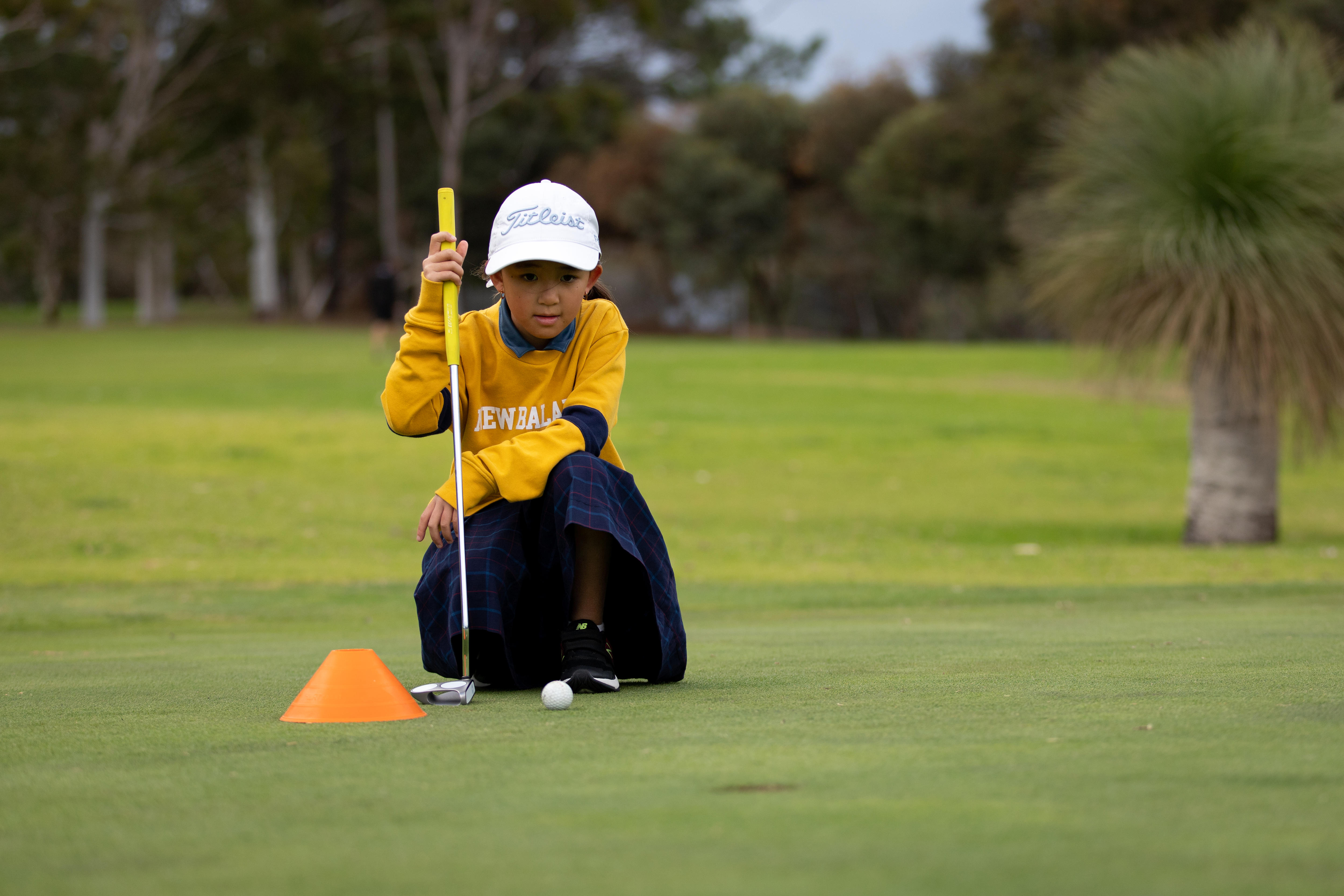 Girl with golf club crouched on her knees on a well-manicured lawn.