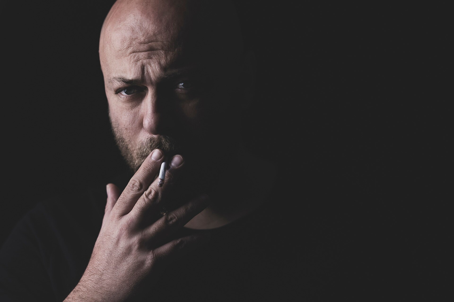 Man smoking a cigarette looking straight ahead with a dark background. 