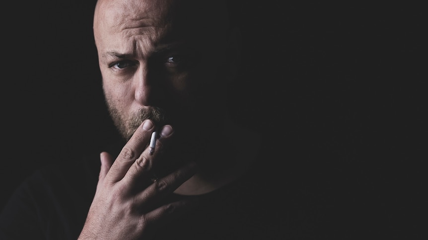 Man smoking a cigarette looking straight ahead with a dark background. 