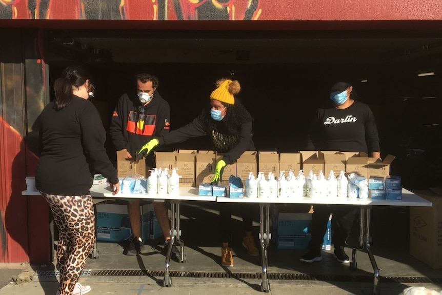 Three VACCHO workers with masks hand out hand sanitiser at a trestle table.