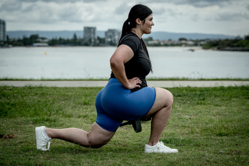 Xanthia lunges with her hand on her hips on a grassy patch by the water outdoors.