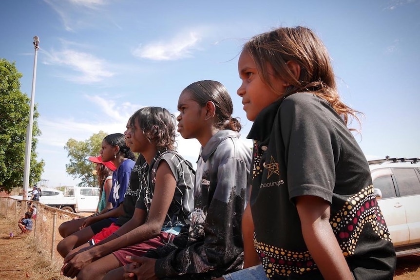 Children sitting on a fence in Bidyadanga at a footy match June 2022. 