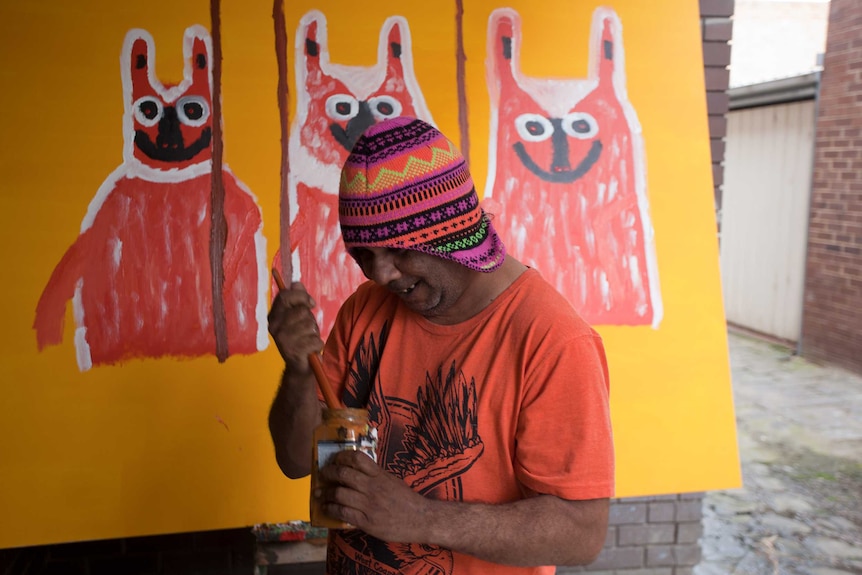 Artist Turbo Brown, holding a jar of paint and paintbrush, in front of a yellow painting of three dogs.
