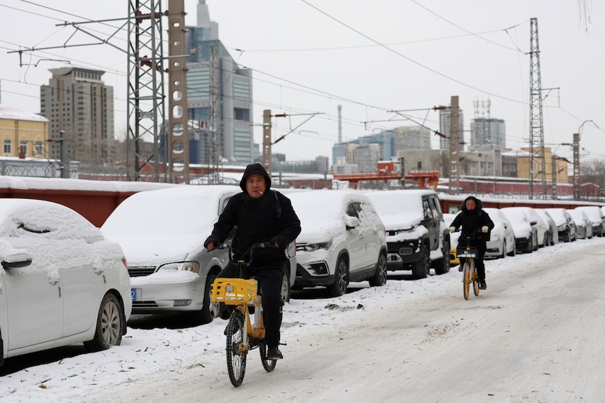 two people ride bicycles on road covered in snow, next to parked cars blanketed by snow