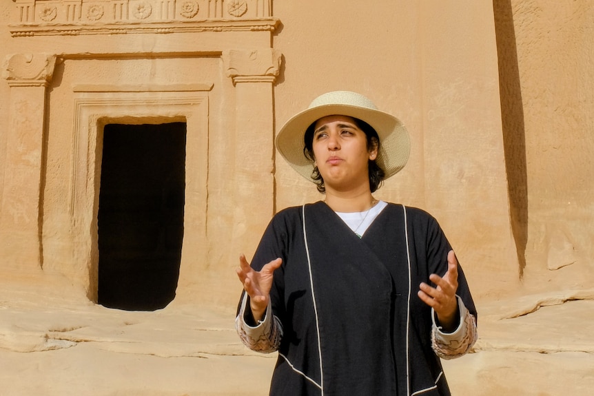 A woman wearing a hat speaks in front of a tomb.