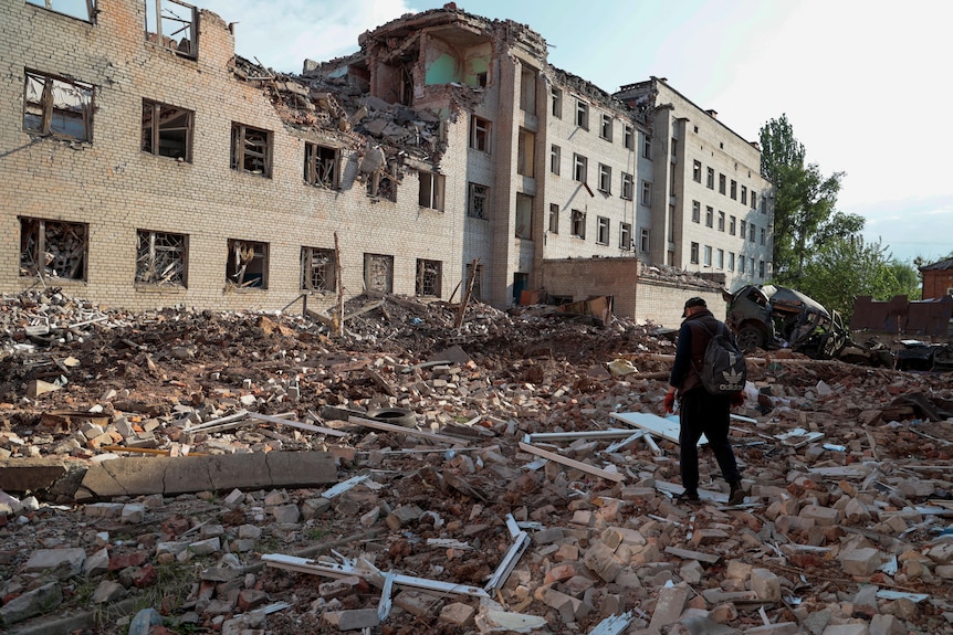 A man walks across rubble next to a building damaged by shelling or rockets. 