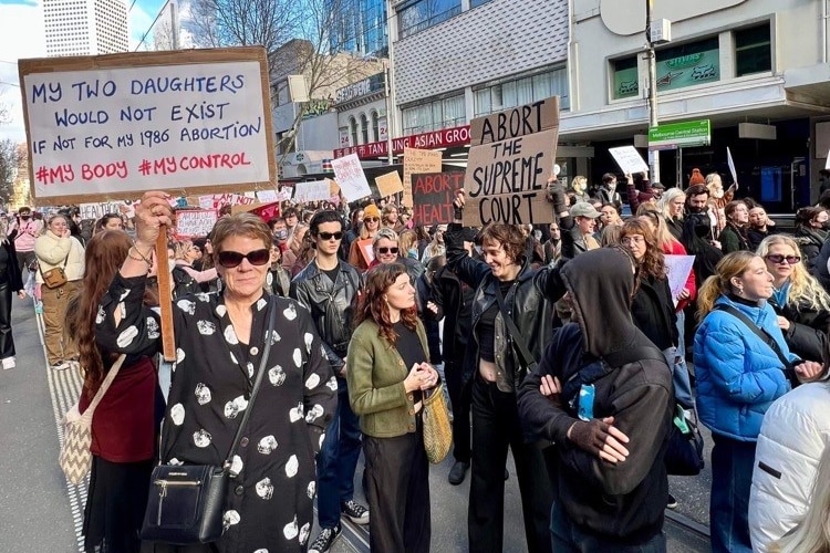 Isla Evans stands in a crowd wearing sunglasses and a spotted dress, and holding a sign.