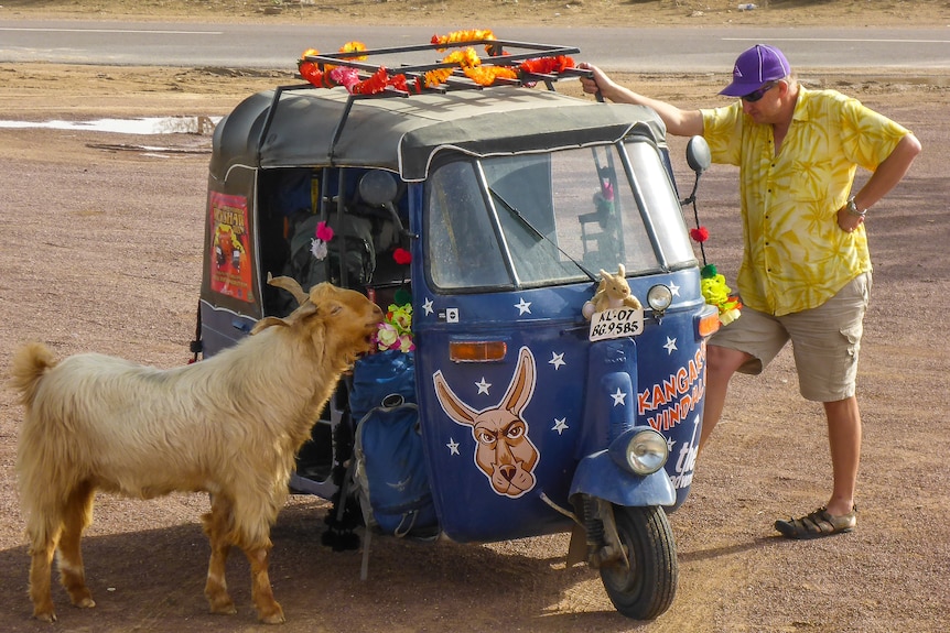 A man stands on one side of a tuk-tuk looking through to a goat on the other side of the vehicle.