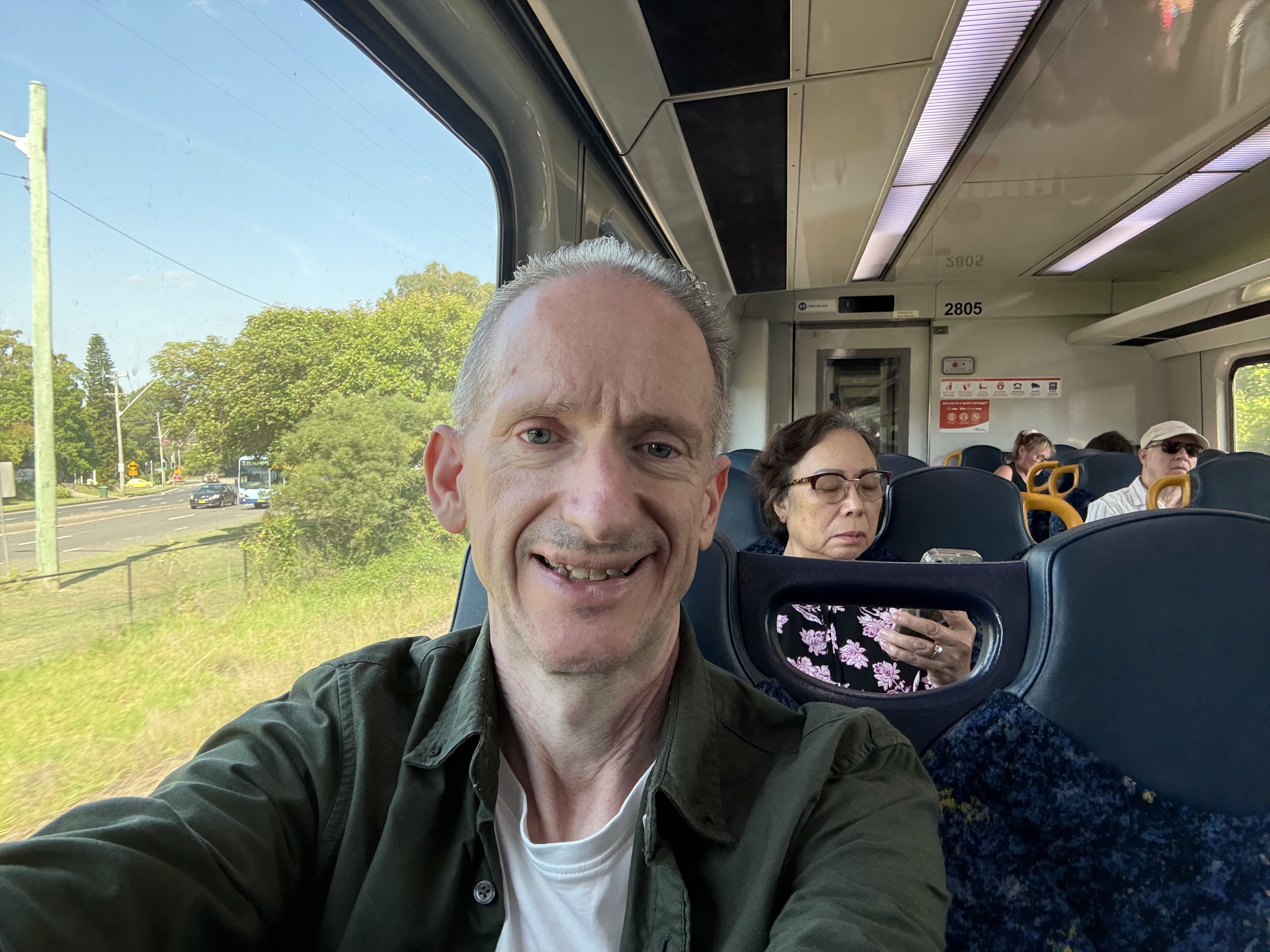 El señor Thomas lleva una chaqueta verde y una camisa blanca y sonríe mientras está sentado en el tren con gente sentada detrás de él.