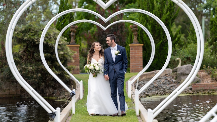 An image of a bride and groom walking through love heart arbors surrounded by a lake and gardens