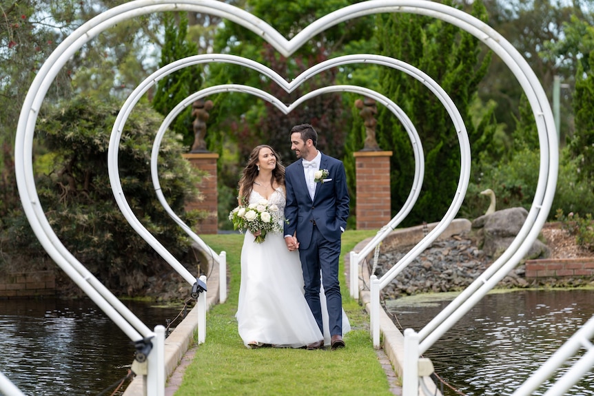 An image of a bride and groom walking through love heart arbors surrounded by a lake and gardens