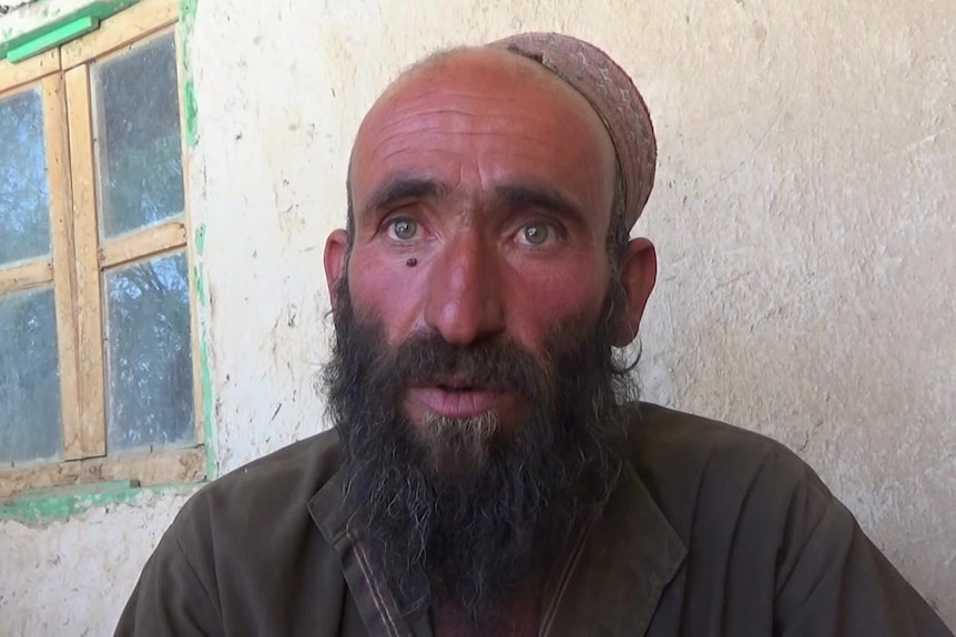 A man with a beard and wearing a cap looks into camera with a serious expression.