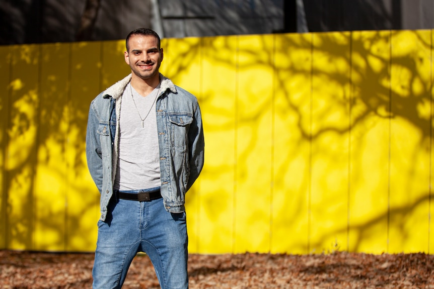 You view a man of Middle Eastern descent standing in a denim jacket and jeans in front of a bright yellow wall.