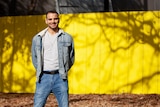 You view a man of Middle Eastern descent standing in a denim jacket and jeans in front of a bright yellow wall.