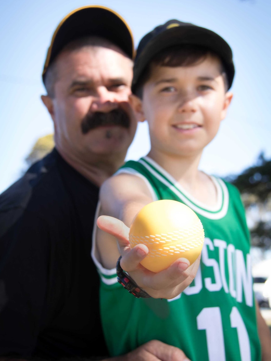 Laurie Marks and his young son Byron, against a brilliant blue sky. Byron holds a bright yellow cricket ball towards the lens.