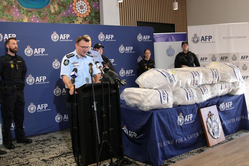 A wide shot of a police media conference with plastic bags filled with cocaine on a table to the right.