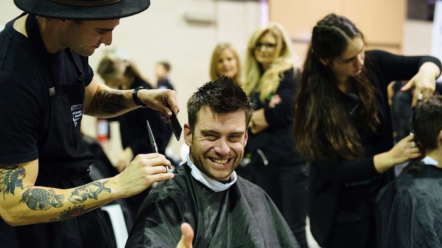 A man with short brown hair receives a haircut from a man with tattooed arms wearing a black felt hat.