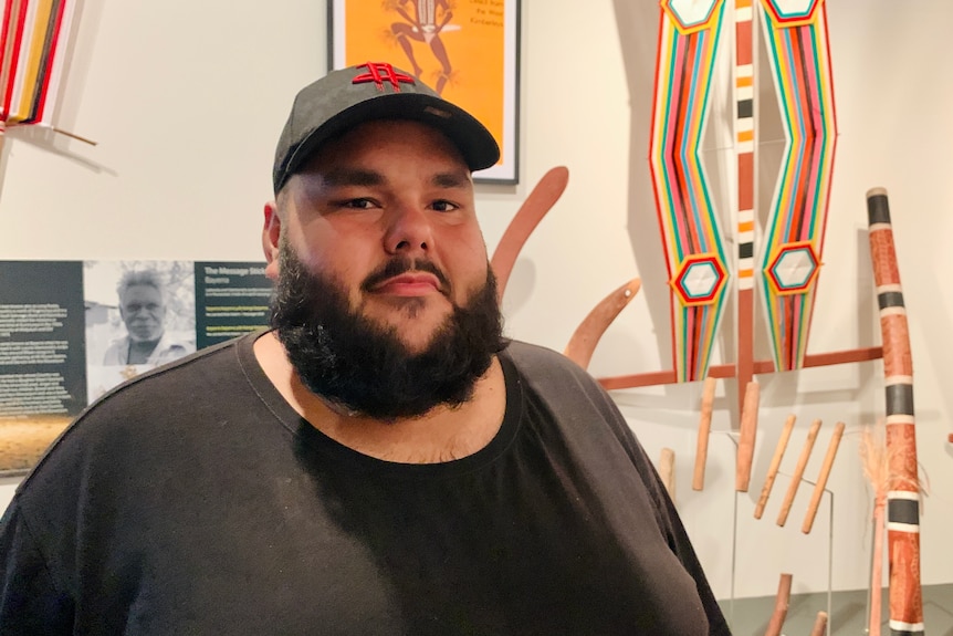 A man in a black shirt and hat stands in front of colourful Aboriginal dance totems